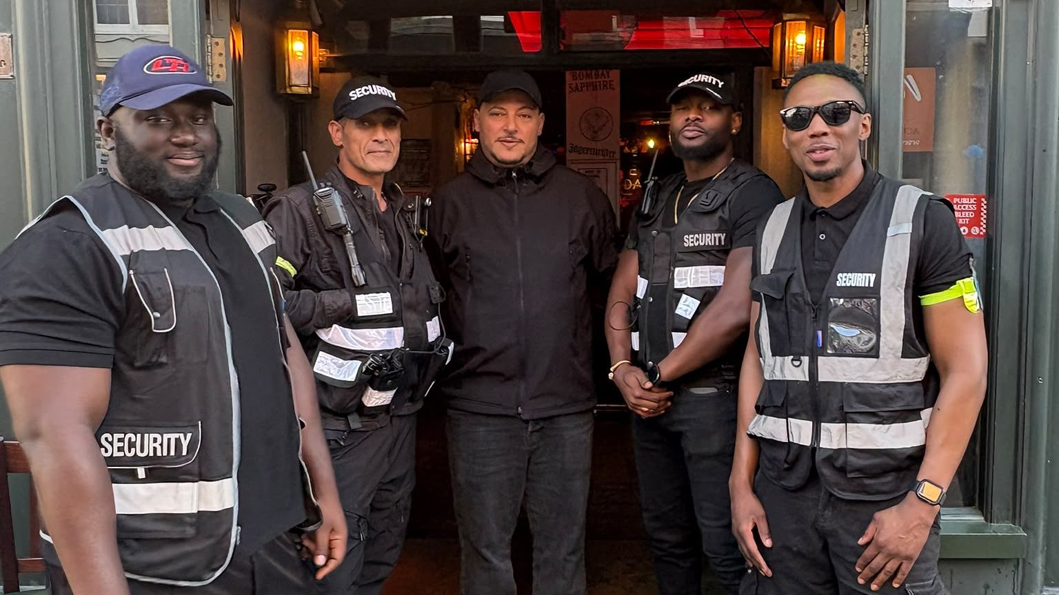 Five licensed door supervisors in high visibility vests standing at the entrance to a London pub.